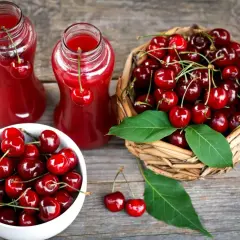Dwa szklane dzbanki z rubinowym sokiem z czereśni, obok koszyk pełen świeżych owoców i miseczka z czereśniami.