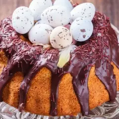 Wilgotna babka gotowana, polana czekoladą i posypana czekoladową posypką, ozdobiona pisankami i piórkiem. Idealna na Wielkanoc.