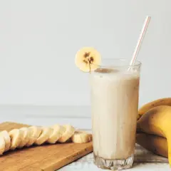 Pyszny koktajl bananowy w szklance, ozdobiony plasterkiem banana. Obok leżą świeże banany i pokrojone na desce.