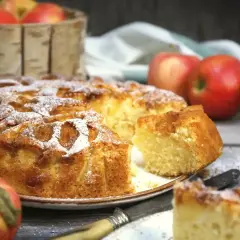 Wilgotne ciasto z jabłkami na oleju, posypane cukrem pudrem, kusi apetycznym wyglądem. Obok koszyk z jabłkami i kilka świeżych owoców.