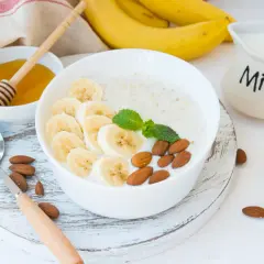 Pyszna owsianka proteinowa z bananem, migdałami i miętą. Obok miseczka z miodem i dzbanuszek z mlekiem.