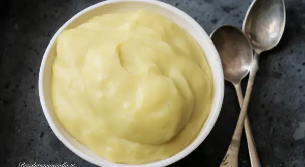 Gładki, żółty krem patissiere w białej miseczce, obok dwie łyżeczki. Idealny do deserów.