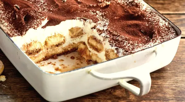 Tiramisu w białej formie, posypane kakao, z widocznymi warstwami biszkoptów i kremu.