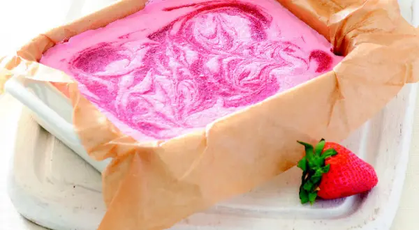 Różowe lody z mascarpone z wirami truskawkowymi w prostokątnym naczyniu wyłożonym papierem.