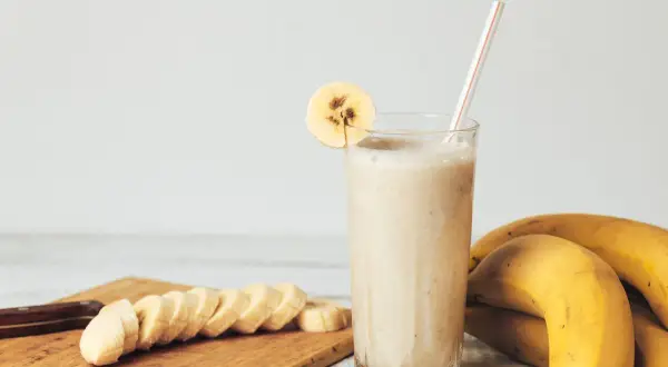 Pyszny koktajl bananowy w szklance, ozdobiony plasterkiem banana. Obok leżą świeże banany i pokrojone na desce.