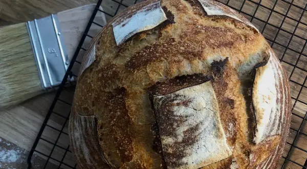 Chleb pszenno żytni na zakwasie, pachnący świeżością, czeka na rozkrojenie. Obok pędzel do smarowania.