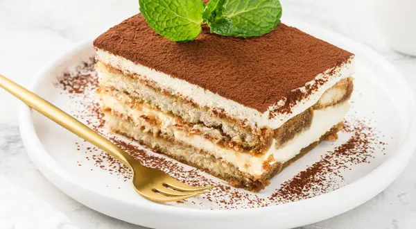Kawałek tiramisu z kremem mascarpone, posypany kakao i udekorowany miętą, na białym talerzu.