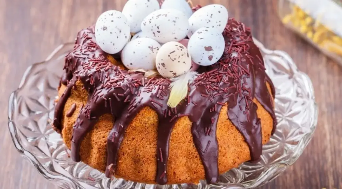 Wilgotna babka gotowana, polana czekoladą i posypana czekoladową posypką, ozdobiona pisankami i piórkiem. Idealna na Wielkanoc.