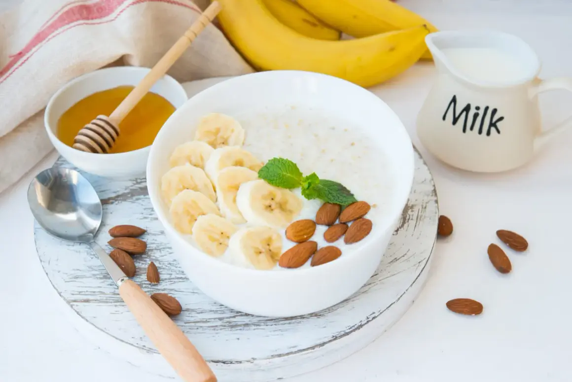 Pyszna owsianka proteinowa z bananem, migdałami i miętą. Obok miseczka z miodem i dzbanuszek z mlekiem.