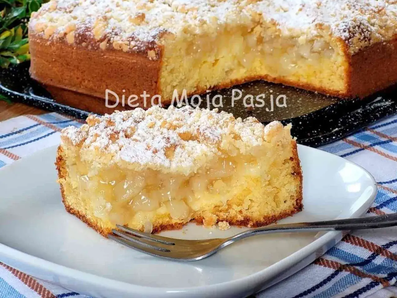 Szybkie tanie ciasto z jabłkami, posypane cukrem pudrem i kruszonką. Pyszny kawałek na talerzyku z widelcem.
