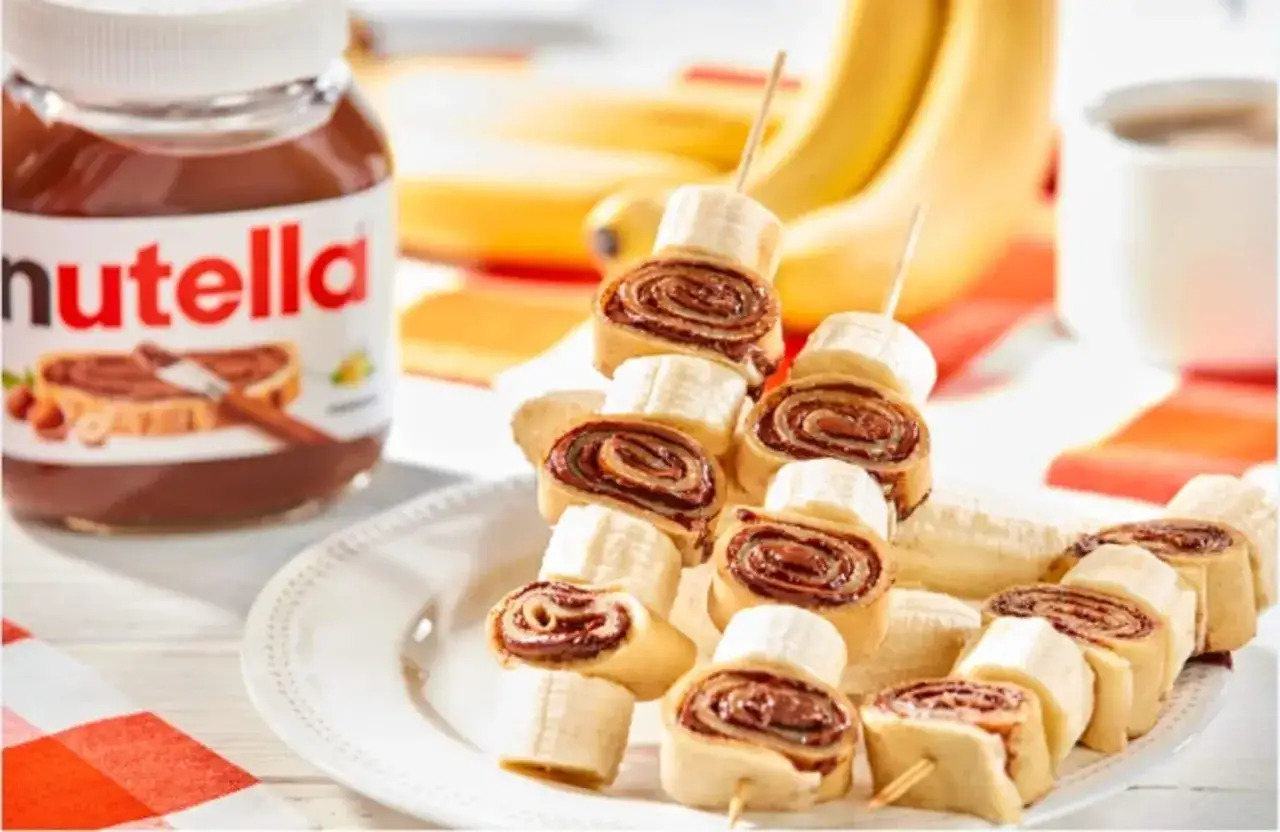 Naleśniki z nutellą i bananem na patyczkach, obok słoik nutelli i banany. Pyszna przekąska!