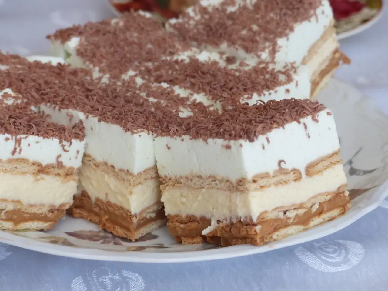Szybkie ciasto bez pieczenia z budyniem i herbatnikami, przełożone kremem karmelowym i posypane wiórkami czekolady.