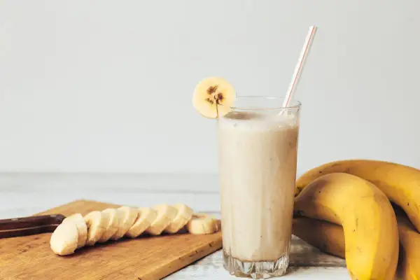 Pyszny koktajl bananowy w szklance, ozdobiony plasterkiem banana. Obok leżą świeże banany i pokrojone na desce.