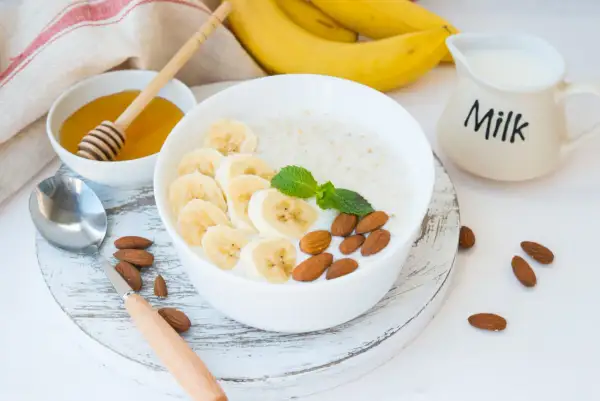 Pyszna owsianka proteinowa z bananem, migdałami i miętą. Obok miseczka z miodem i dzbanuszek z mlekiem.